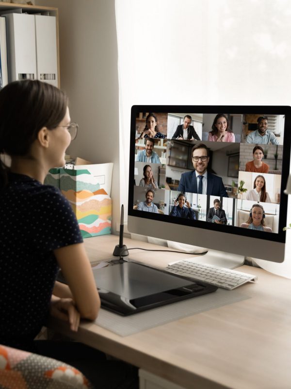 Happy millennial female employee holding video call negotiations meeting. Back rear view happy millennial female employee looking at computer monitor, holding online video call negotiations meeting with diverse colleagues and male executive manager, working from home.